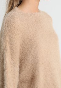 Lichtbeige, fluffy gebreide trui met een wijde halslijn, losse pasvorm en zachte textuur, die de gezellige uitstraling en eenvoud benadrukt.