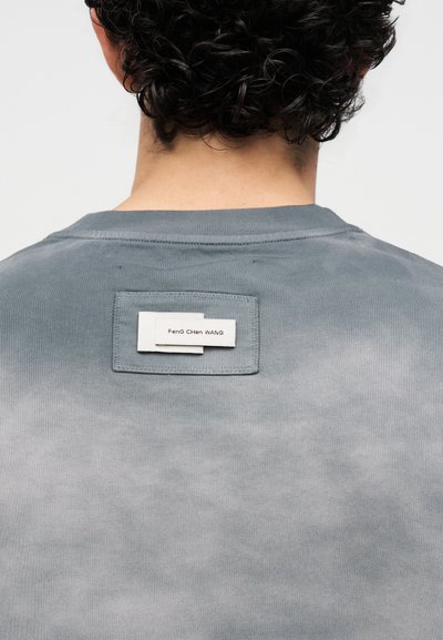 Άτομο με σγουρά μαύρα μαλλιά, που βλέπουμε από πίσω, φοράει γκρι πουκάμισο με ένα ραμμένο ορθογώνιο ταμπελάκι με όνομα στο πάνω μέρος της πλάτης.