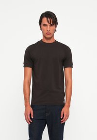 Ung mand med mørkt hår iført en ensfarvet sort T-shirt med korte ærmer og mørkeblå jeans, der står mod en ensfarvet hvid baggrund.