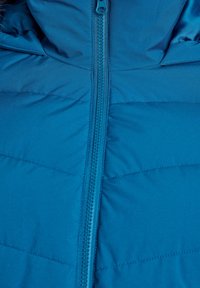 Veste matelassée bleue avec fermeture éclair centrale et coutures visibles sur le tissu, montrant un gros plan de la section avant supérieure.