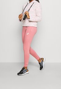 Helle rosa, taillierte Jogginghosen mit einem weißen Logo, kombiniert mit einem gestreiften, rosa und weißen Pullover mit Kapuze, dazu schwarze Sportschuhe.