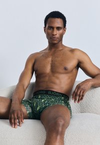 Zeleni boksarji z vzorcem in črnim pasom z napisom "BJÖRN BORG". Model sedi na teksturirani svetli površini.