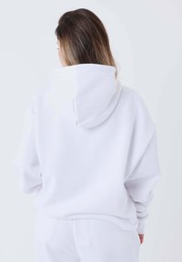 Sudadera blanca de gran tamaño con una capucha amplia, hombros caídos y una tela suave y texturizada. Las costuras se extienden para un ajuste relajado.