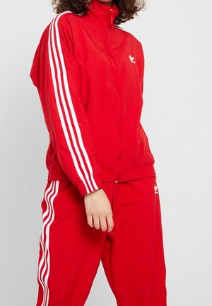 Persoon draagt een rood Adidas trainingspak met witte strepen op de mouwen en broek, staand tegen een effen lichte achtergrond.