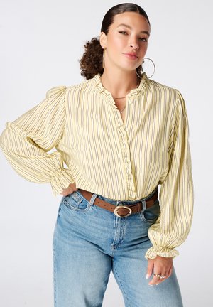 Vrouw met krullend haar in een lage paardenstaart draagt grote creolen, een geel gestreept bloesje met ruches aan kraag en manchetten, een blauwe spijkerbroek en een bruine riem.
