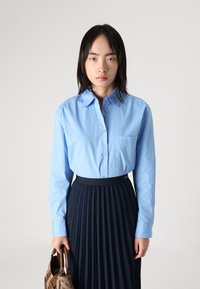 Light blue button-up shirt with a chest pocket, paired with a pleated navy skirt. The individual holds a brown patterned handbag.