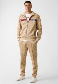 Beige trainingsjack en broek set met een herhalend logopatroon, rode en zwarte accentstrepen, ritsluiting, geribbelde manchetten, gecombineerd met witte sneakers.