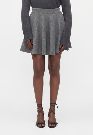 Femme portant un haut à manches longues gris foncé, une jupe plissée grise et des sandales noires à talons hauts à brides, debout devant un fond uni.