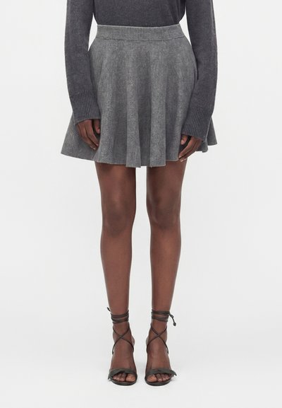 Femme portant un haut à manches longues gris foncé, une jupe plissée grise et des sandales noires à talons hauts à brides, debout devant un fond uni.