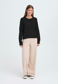 Pull en maille noir avec col rond, associé à un pantalon beige à jambes larges et des baskets noires. L'ensemble a une coupe décontractée et un tissu texturé.