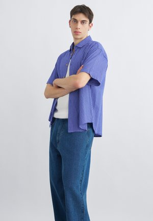 Ung mand stående med armene krydset iført en blåternet kortærmet skjorte, hvid t-shirt og blå jeans mod en ensartet lys baggrund.
