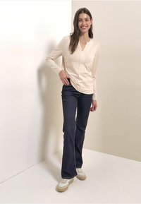 Cremefarbenes Langarmoberteil mit V-Ausschnitt, kombiniert mit dunkelblauen Schlagjeans und hellfarbenen Sneakers. Glatter Stoff, taillierte Passform.