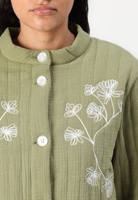 Veste matelassée vert olive avec un col rond, ornée de broderies florales blanches et de quatre boutons sur le devant.