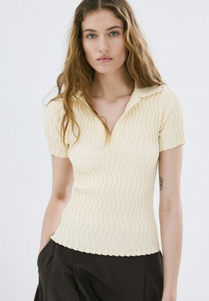 Joven con cabello largo y ondulado, vistiendo una camiseta corta de manga corta en color crema con textura de canalé y pantalones negros, de pie contra un fondo blanco liso.