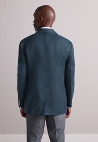 Türkisfarbener Leinenblazer mit glatter Textur, mit Kerbkragen und silbernen Knopfakzenten, kombiniert mit grauer Hose. Ansicht von hinten.