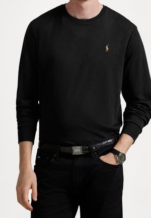 Mies, jolla on päällään istuva musta pitkähihainen paita, jossa on pieni kirjailtu polo-pelaajan logo, mustat housut, musta vyö ja musta rannekello.