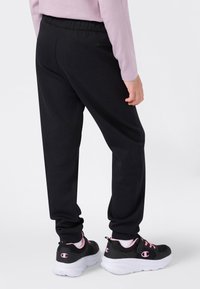 Person in schwarzer Jogginghose und schwarzen Sneakers mit pinken Akzenten auf weißen Sohlen, steht vor einem schlichten weißen Hintergrund.