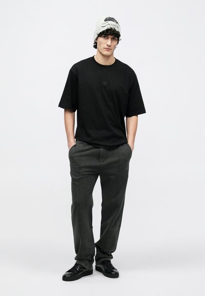 Black t-shirt with a subtle logo, paired with dark gray pants featuring side stitching details; black lace-up shoes; light gray beanie.