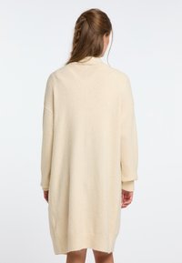 Knee ilgio, kreminės spalvos megztinis su laisvu siluetu, žemais pečiais ir glotniais rankogaliais. Minkšta tekstūra, minimalus detalių kiekis ir vientisas dizainas.