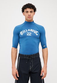 Zils īsām piedurknēm un augstu apkakli ar balto "BILLABONG" logo; kopā ar melniem peldšortiem ar auklu viduklī.