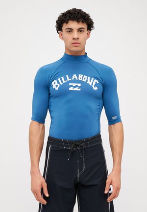 Blå kortermet rashguard med høy krage og hvit "BILLABONG"-logo; kombinert med svarte svømmetober med snor i livet.