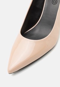Gros plan d'une chaussure à talon haut en cuir verni beige à bout pointu avec une doublure intérieure noire sur fond blanc.