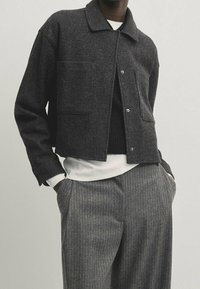 Donkergrijze wollen jas met een kraag, twee voorzakken en drukknoopsluiting, gedragen over een zwart vest en een wit overhemd.