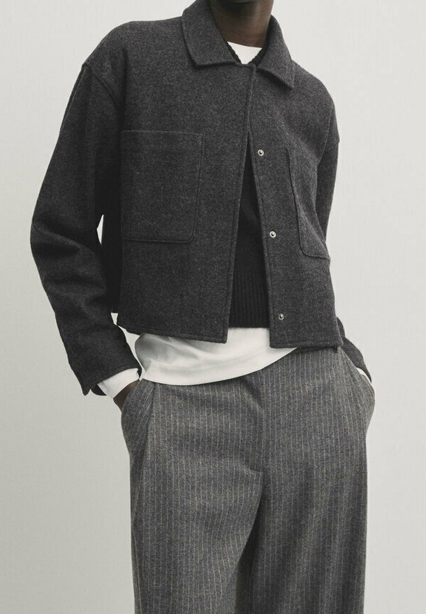 Donkergrijze wollen jas met een kraag, twee voorzakken en drukknoopsluiting, gedragen over een zwart vest en een wit overhemd.