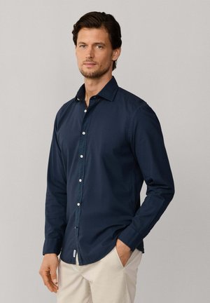 Homme debout avec une main dans la poche, portant une chemise boutonnée bleu foncé et un pantalon beige clair sur un fond uni.
