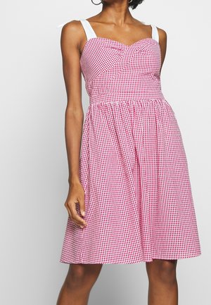 Kvinde iført ærmeløs rød og hvidternet kjole med hvide bindebånd og indsamlet nederdel, stående mod en ensfarvet baggrund.