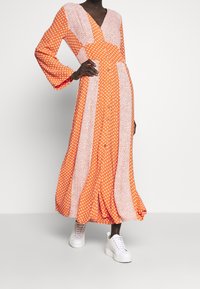 Robe longue orange à motifs avec manches évasées, ornée de pois blancs et de sections florales, fermeture à boutons, portée avec des baskets blanches.