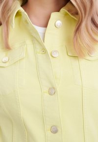 Giacca di jeans giallo con bottoni a pressione argentati, due tasche sul petto e un colletto classico. Tessuto testurizzato con dettagli di cucitura visibili.