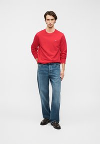 Sweat-shirt rouge à col rond avec manches longues, associé à un jean droit bleu clair. Des chaussures marron complètent cette tenue décontractée.