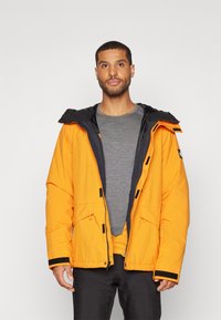 Oranž puffer-jakk musta kapuutsiga, millel on tõmbluku ja nööpide kinnitused, esiõhukotid ja mustad klambrid. All on hall särk.