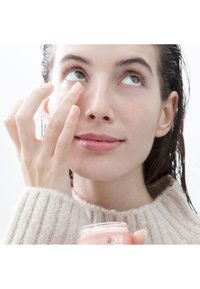 Crème hydratante dans un pot en verre transparent avec un couvercle couleur pêche, tenue par une personne appliquant de la crème sous son œil. Le fond est blanc uni.