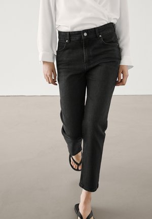 Woman wearing black straight-leg jeans, white long-sleeve blouse, and black open-toe sandals walking forward on light floor.