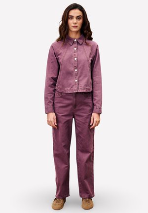 Femme aux longs cheveux bruns portant une veste et un pantalon en denim violet assortis, se tenant droite avec une expression neutre et des chaussures marron.