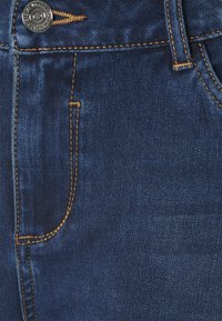 Primer plano de unos pantalones vaqueros de denim azul oscuro que muestran un botón de metal, costura amarilla, bolsillo delantero y presilla para el cinturón.