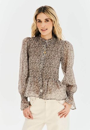 Kobieta w długim rękawie, zapinanej na guziki kwiatowej bluzce z bufkami na ramionach oraz jasnych spodniach, stojąca na jednolitym tle.