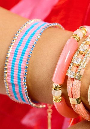 Des bracelets colorés sur un poignet comprennent un bracelet rayé rose et bleu, un bracelet de perles roses lisses, et un design tressé texturé avec des accents.
