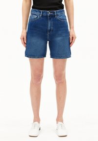 Shorts en denim taille haute, en bleu moyen. Présentent cinq poches, des passants de ceinture et un ourlet brut. Associés à des chaussures blanches structurées.