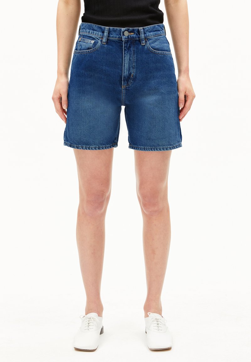 Shorts en denim taille haute, en bleu moyen. Présentent cinq poches, des passants de ceinture et un ourlet brut. Associés à des chaussures blanches structurées.
