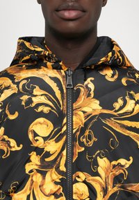 Närbild av en person som bär en svart hooded jacka med ett gyllene barockblommönster och en central dragkedja.