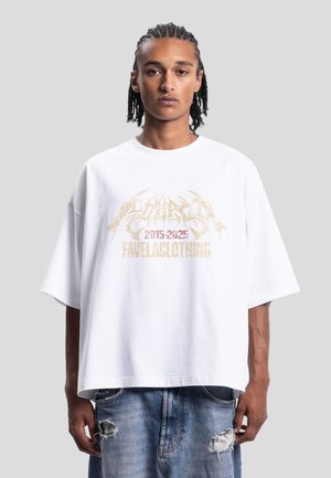 Jeune homme aux cheveux tressés portant un t-shirt blanc oversize avec un texte doré "2015-2025 Favelaclothing" et un jean bleu déchiré.