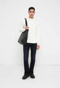 Uomo che indossa un maglione bianco, jeans blu scuro e scarpe nere, mentre tiene una grande borsa a tracolla in pelle nera sulla spalla, su uno sfondo neutro.