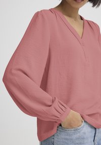 Rosa bluse med V-hals og knapdetalje, der har lange, puffede ærmer og et tekstureret, let stof.