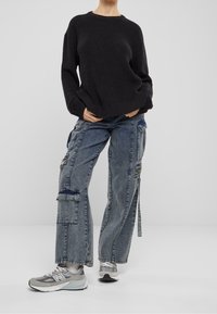 Osoba w luźnym czarnym swetrze, szerokich niebieskich dżinsach cargo z kieszeniami i szarych trampkach New Balance, stojąca na jednolitym tle.