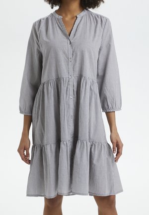 Femme portant une robe grise à manches longues, boutonnée à l'avant avec des rayures verticales fines et une jupe à volants froncée.