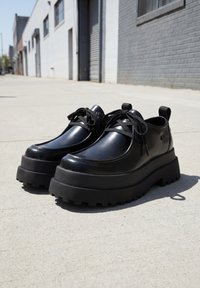 Ζευγάρι μαύρα chunky παπούτσια σε πλατφόρμα με κορδόνια τοποθετημένα σε πεζοδρόμιο από σκυρόδεμα με ηλιακό φως κοντά σε γκρι και λευκό κτίριο από τούβλα.