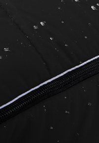 Schwarzer wasserdichter Stoff mit strukturierter Oberfläche, versehen mit Wassertropfen und einem kontrastierenden Reißverschluss mit weißen Akzenten am Rand.